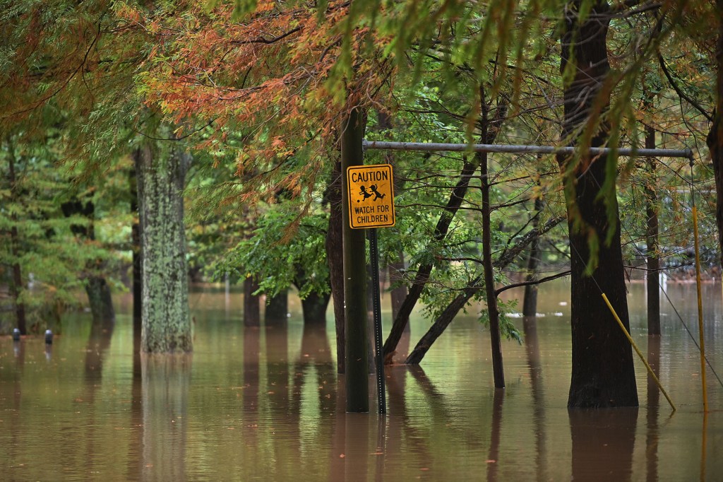 A yellow “Caution: Watch for Children” sign stands partially submerged in floodwater among trees, with water covering the ground beneath it.