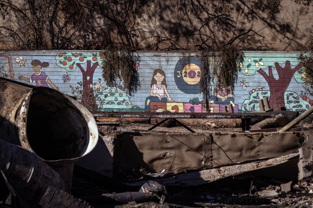 A painted mural depicting children playing is visible on a wall behind piles of broken concrete, pipes, and debris in a fire damaged outdoor playground.