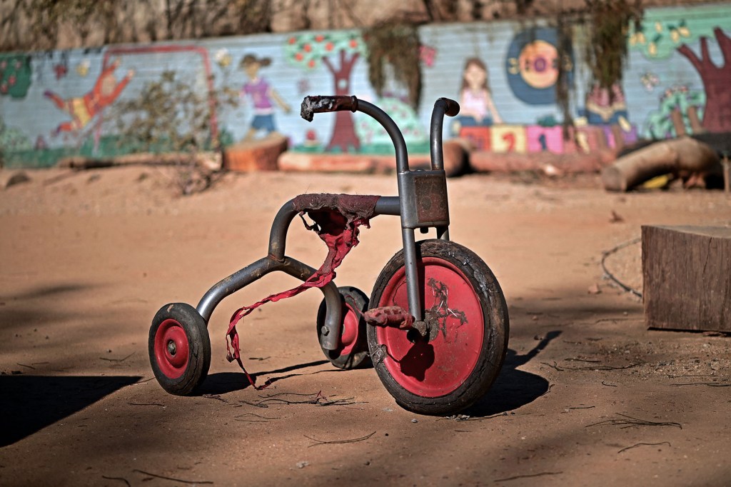 small red tricycle with torn fabric and damaged wheels sits on dusty ground, with a children’s mural blurred in the background.