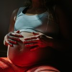 A woman holds her pregnant belly in the shadows.