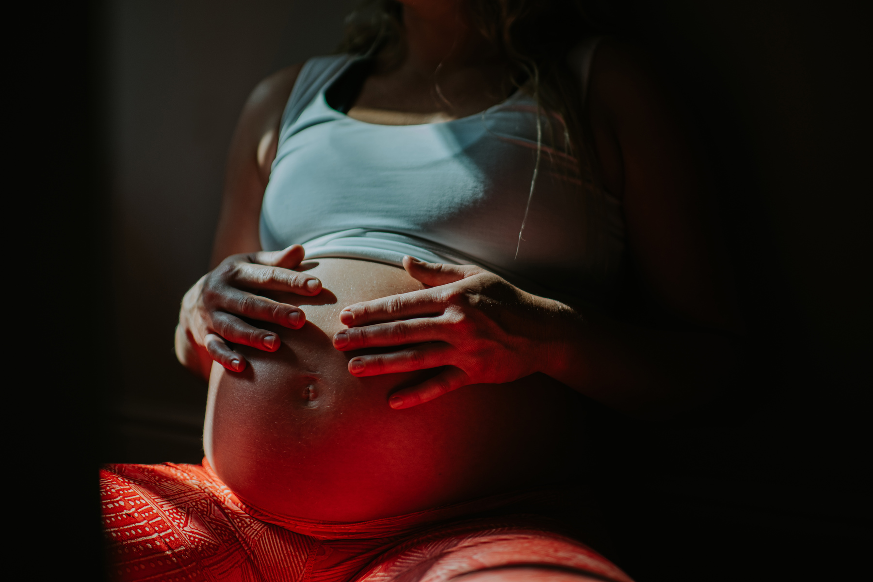 A woman holds her pregnant belly in the shadows.