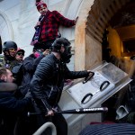a crowd of Trump supporters in helmets and gas masks presses into a boarded-up arched entrance of the U.S. Capitol in Washington, D.C., as one swings a baseball bat and another carries a clear riot shield toward police blocking the hallway.