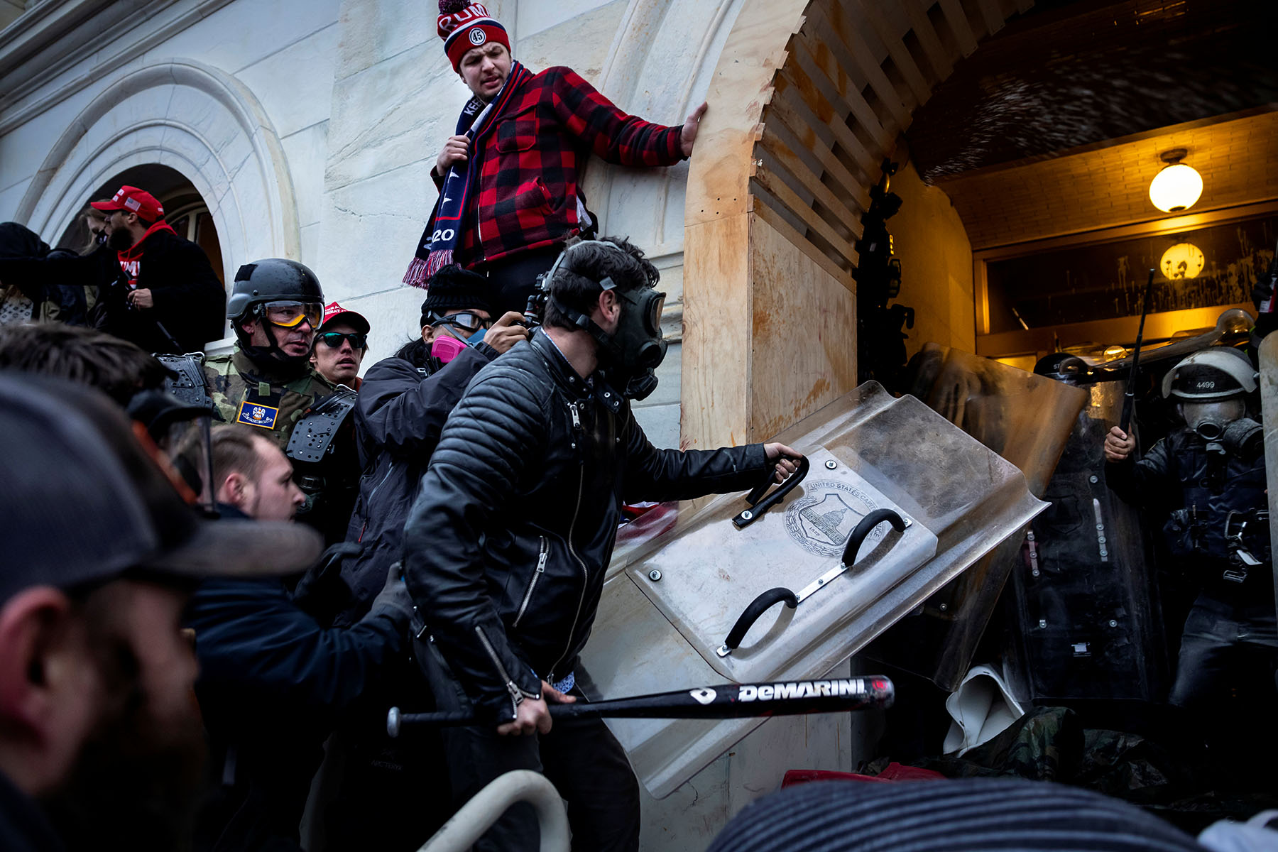 a crowd of Trump supporters in helmets and gas masks presses into a boarded-up arched entrance of the U.S. Capitol in Washington, D.C., as one swings a baseball bat and another carries a clear riot shield toward police blocking the hallway.