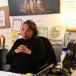 A woman sits at a messy desk with her hands folded, listening.