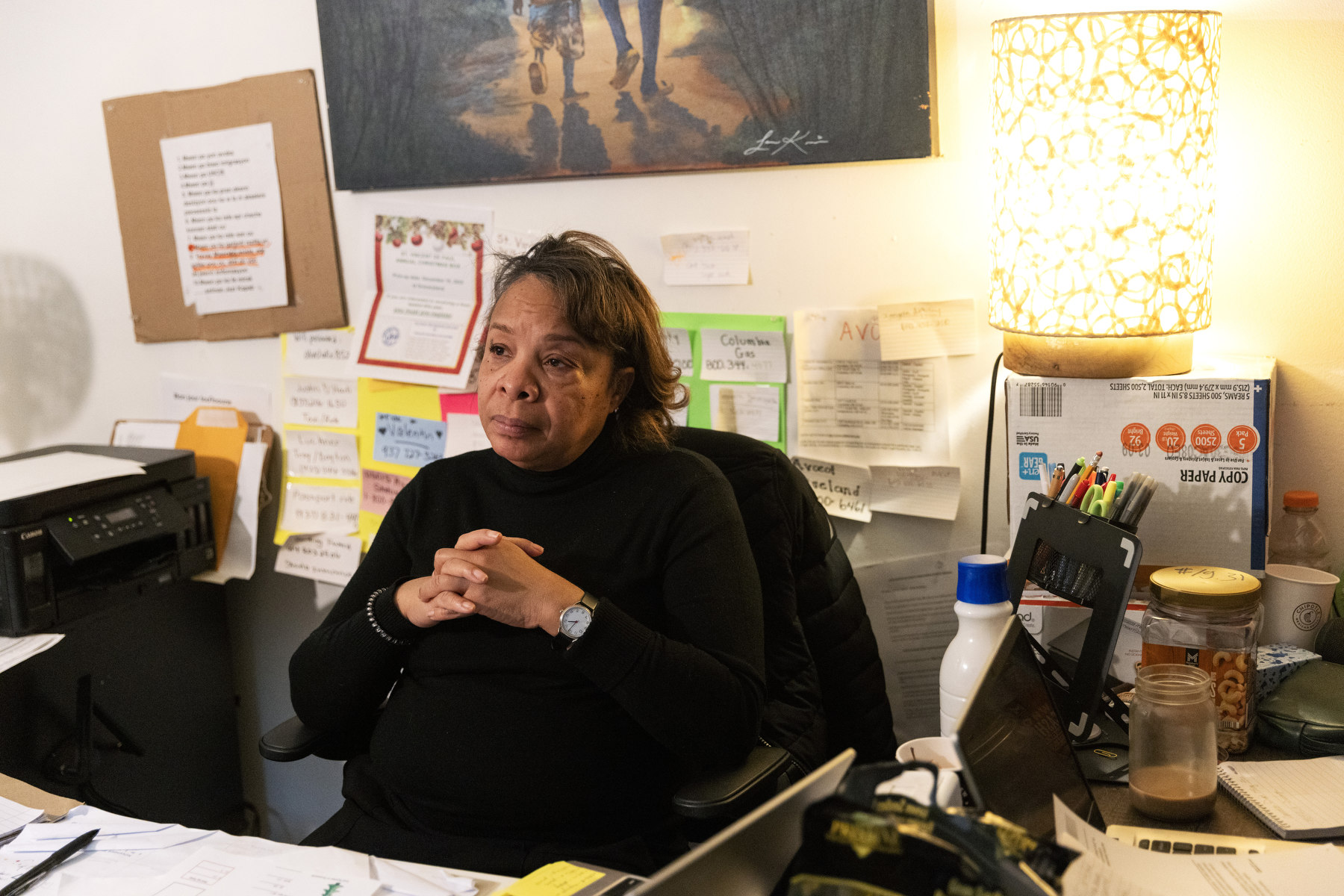 A woman sits at a messy desk with her hands folded, listening.