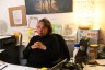 A woman sits at a messy desk with her hands folded, listening.