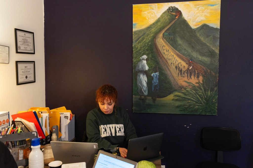 A woman sits at a desk with a painting behind her.