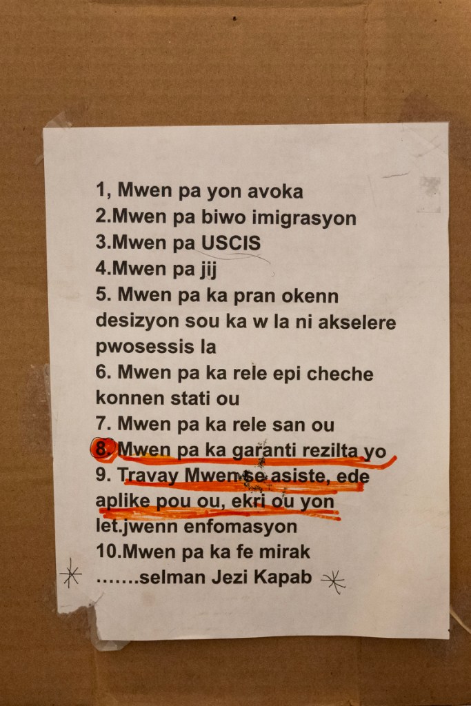A white sign with Haitian Creole writing is taped to cardboard on a wall.
