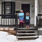 Two children hold protest signs on a front stoop surrounded by snow.
