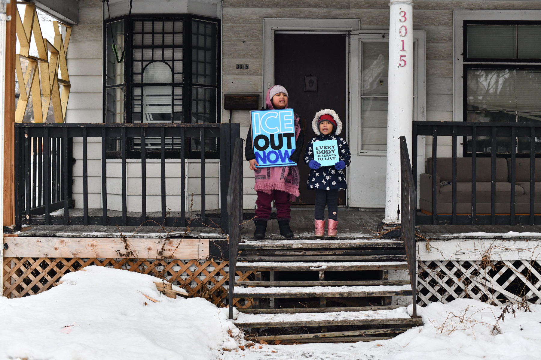Two children hold protest signs on a front stoop surrounded by snow.