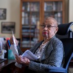 Norton sits in a chair in an office holding papers as she looks toward the camera.