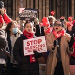A crowd of nurses and supporters wearing red union clothing chant and hold signs reading “Stop Silencing Nurses” and “Cientes antes que ganancias” during a strike outside a hospital.