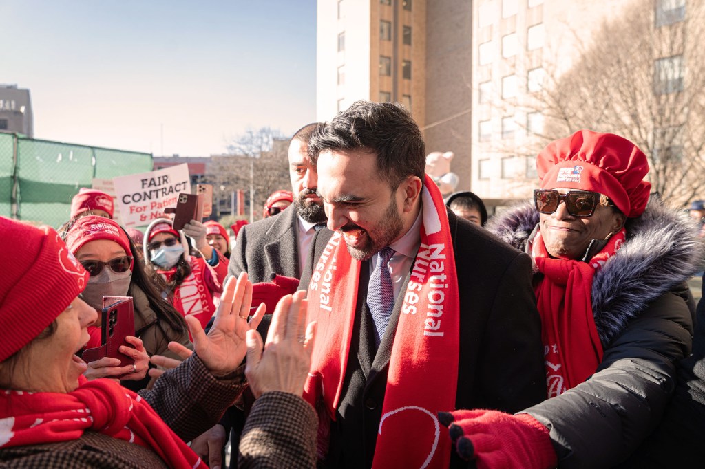 New York City Mayor Zohran Mamdani wearing a red scarf greets and high-fives striking nurses bundled in winter coats and red union gear outside a hospital on a sunny winter day.