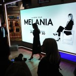 Melania walks in silhouette past a billboard advertising her film while surrounded by other silhouettes.