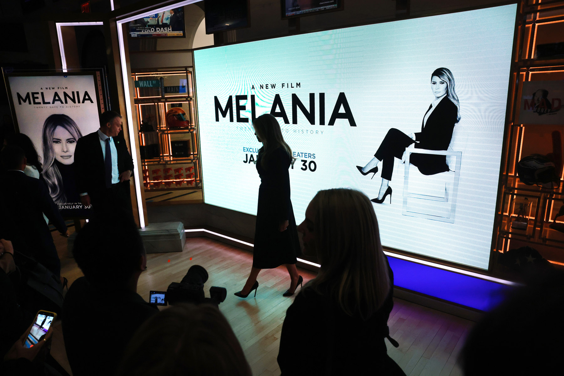 Melania walks in silhouette past a billboard advertising her film while surrounded by other silhouettes.