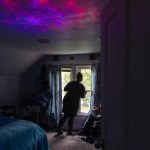 A woman use seen from behind, looking out her window in a darkened room.