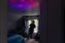 A woman use seen from behind, looking out her window in a darkened room.