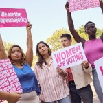 A group of women stand holding bright pink protest signs that read “women are voting” and “we are the supermajority,” under a clear sky.