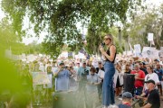 Dr. Annie Andrews speaks into a microphone at an outdoor campaign event, standing before a large crowd holding signs.