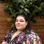 A woman in a colorful floral dress looks at the camera surrounded by greenery and a wooden wall.