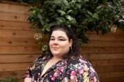 A woman in a colorful floral dress looks at the camera surrounded by greenery and a wooden wall.
