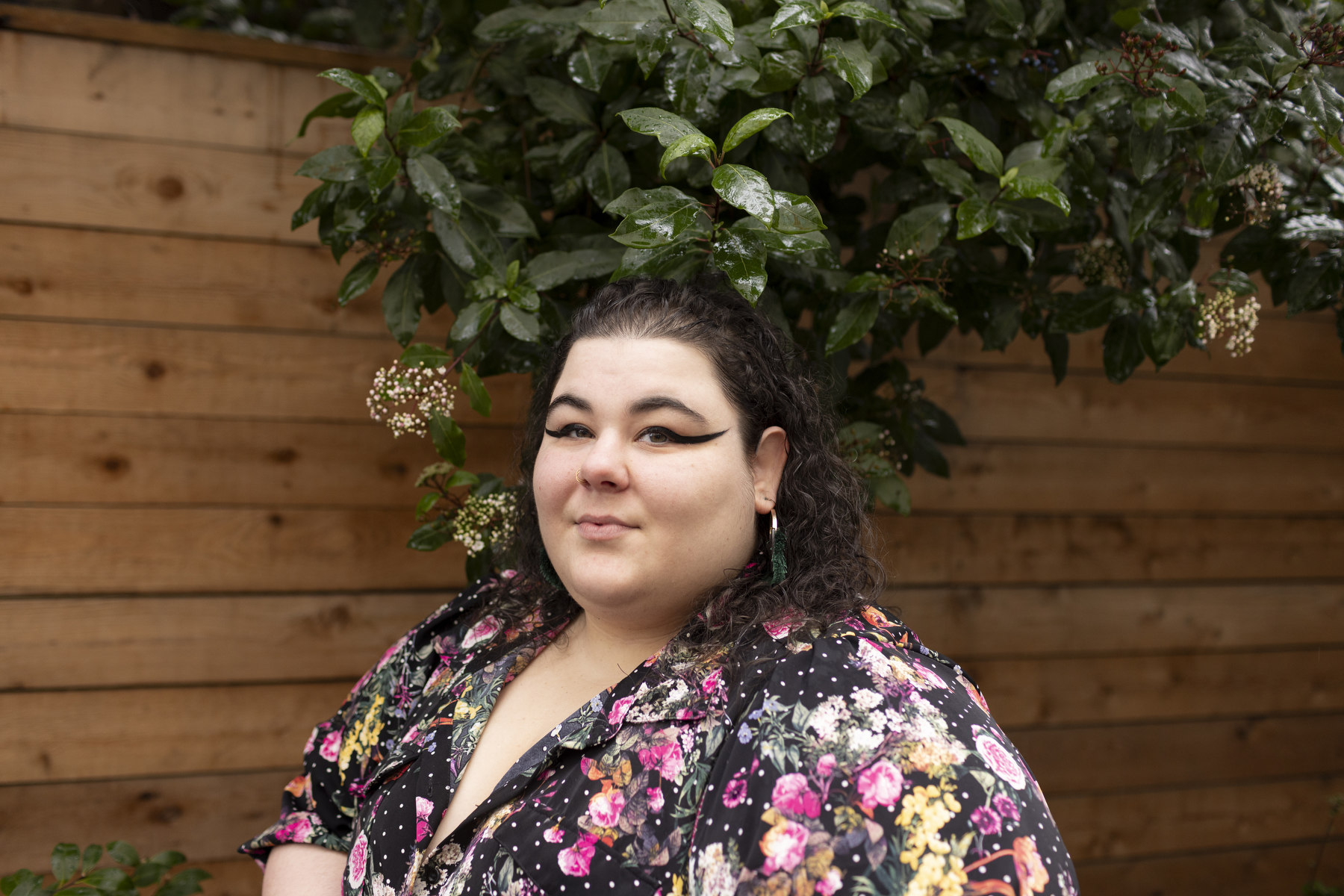 A woman in a colorful floral dress looks at the camera surrounded by greenery and a wooden wall.