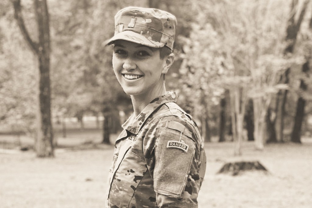 Mikayla Blaska smiles while wearing a U.S. Army uniform and patrol cap during Ranger School training, standing outdoors among trees.