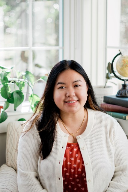 Brittany Vang sits in a bright room near a window with houseplants and books behind her. She smiles at the camera, wearing a white cardigan over a patterned red top and a gold necklace.