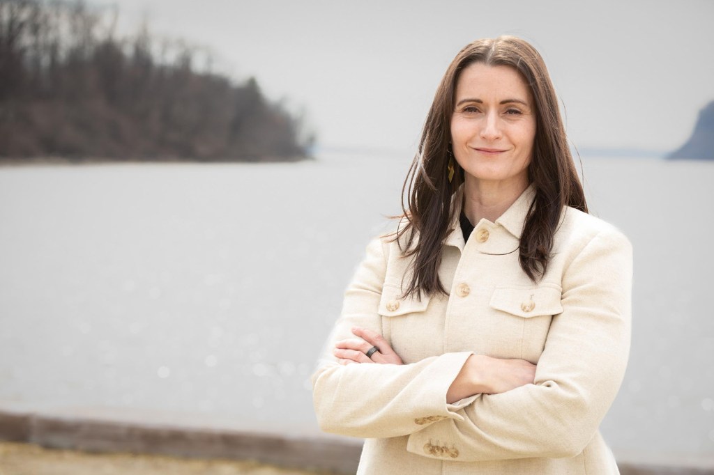 Cait Conley stands outdoors near a body of water with her arms crossed, wearing a light-colored jacket.