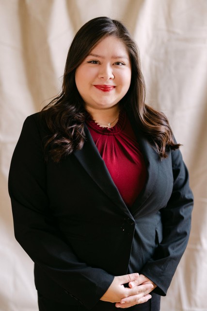 Chloe Urias poses indoors against a softly lit neutral backdrop, wearing a black blazer over a red blouse. She faces the camera with a slight smile, her dark hair styled in loose waves.