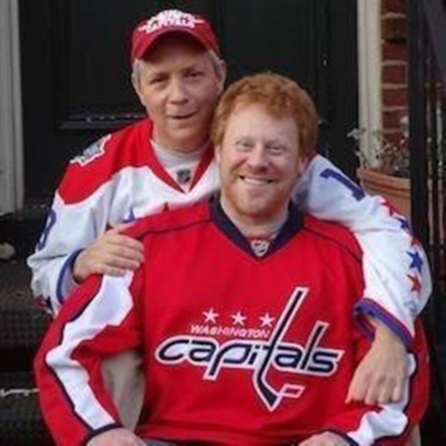Two men stand close together outdoors wearing Washington Capitals hockey jerseys, smiling at the camera with one man’s arm around the other.