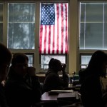 Silhouetted high school students sit through class under a U.S. flag hanging on a window.