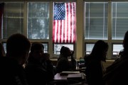 Silhouetted high school students sit through class under a U.S. flag hanging on a window.