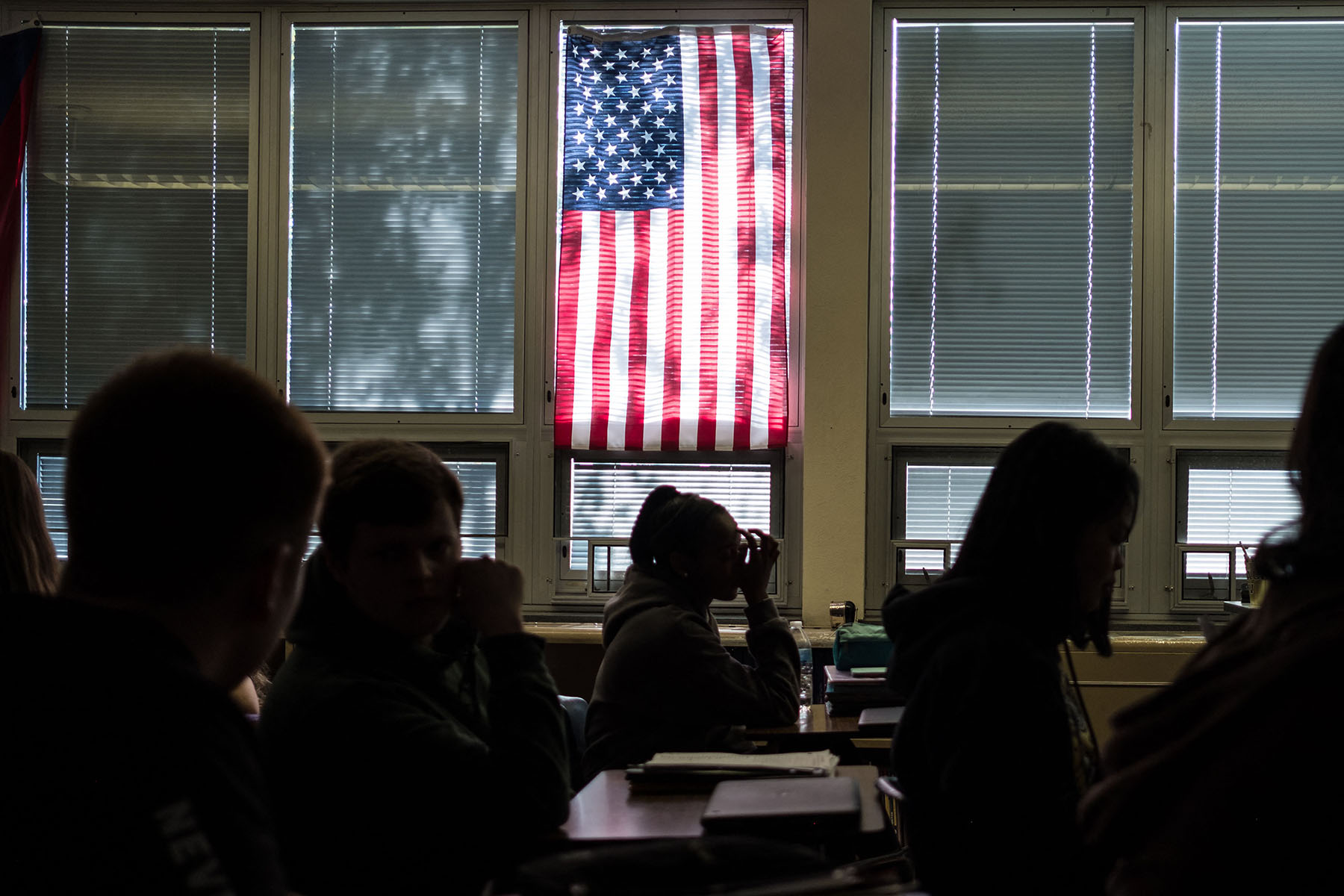 Silhouetted high school students sit through class under a U.S. flag hanging on a window.