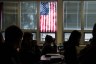 Silhouetted high school students sit through class under a U.S. flag hanging on a window.