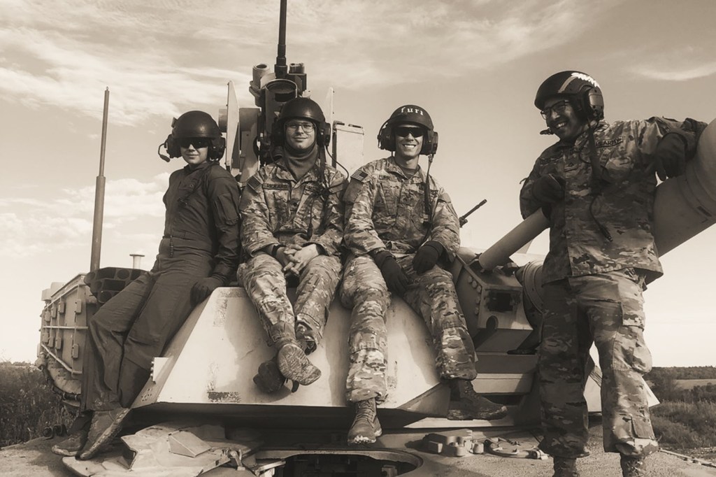 Elizabeth Dempsey Beggs stands with three fellow soldiers seated on a military vehicle, all wearing helmets and Army uniforms during training.