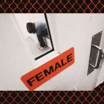 Photo illustration showing a woman hunched on a bench inside an ICE detention facility, seen through a small window in a heavy white door labeled “female,” with an orange chain-link fence pattern framing the image.