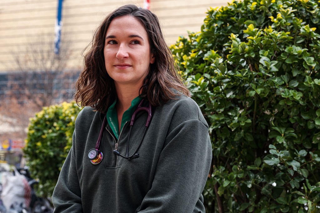 A doctor wearing a green fleece and a stethoscope stands outdoors near bushes, looking slightly off camera.