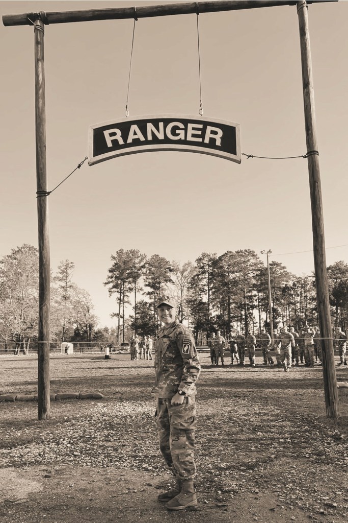 Sara Febbo stands beneath a wooden sign reading “RANGER” while wearing a U.S. Army uniform, with soldiers lined up in the background.