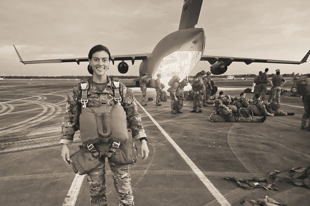Hailey Gibbons stands on an airfield in front of a military aircraft while wearing a parachute harness and U.S. Army uniform, with soldiers preparing equipment behind her.