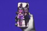 A hand holds a smartphone against a solid purple background. On the phone’s screen, a black-and-white image shows two officers walking away, wearing vests labeled “POLICE ICE,” partially obscured by pink digital glitch lines.