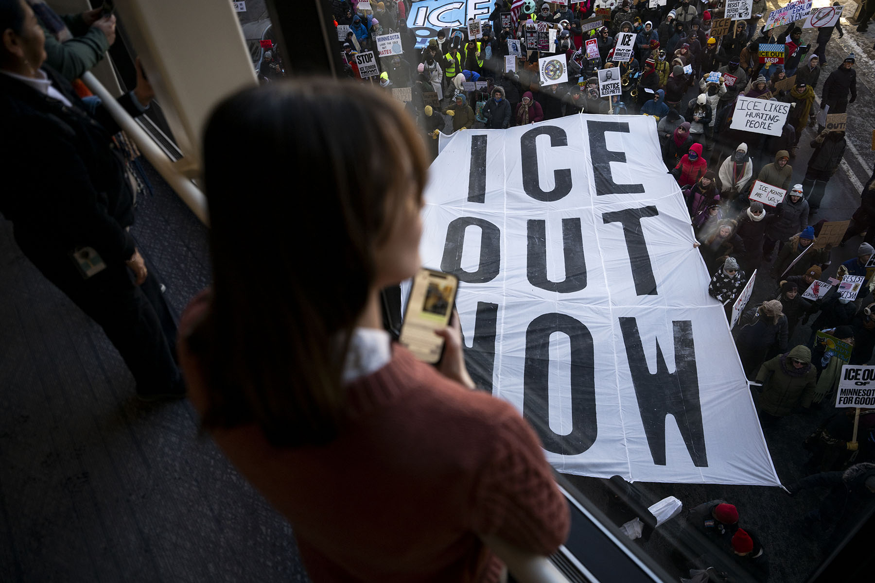 People look on from a skyway as demonstrators march during a protest against ICE enforcement carrying a large banner that reads "ICE OUT NOW."