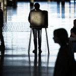 A voter stands at a ballot booth inside a large, dimly lit polling place, silhouetted against bright light from the entrance, while poll workers and other voters move nearby.