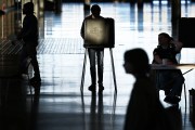 A voter stands at a ballot booth inside a large, dimly lit polling place, silhouetted against bright light from the entrance, while poll workers and other voters move nearby.