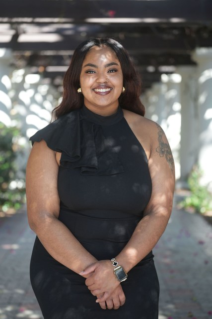 Shawnie Allen stands outdoors in dappled sunlight, smiling at the camera in a sleeveless black dress with a ruffled shoulder detail.