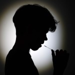 A silhouetted child holds a thin swab to their mouth, while taking an oral COVID-19 test against a softly lit background.