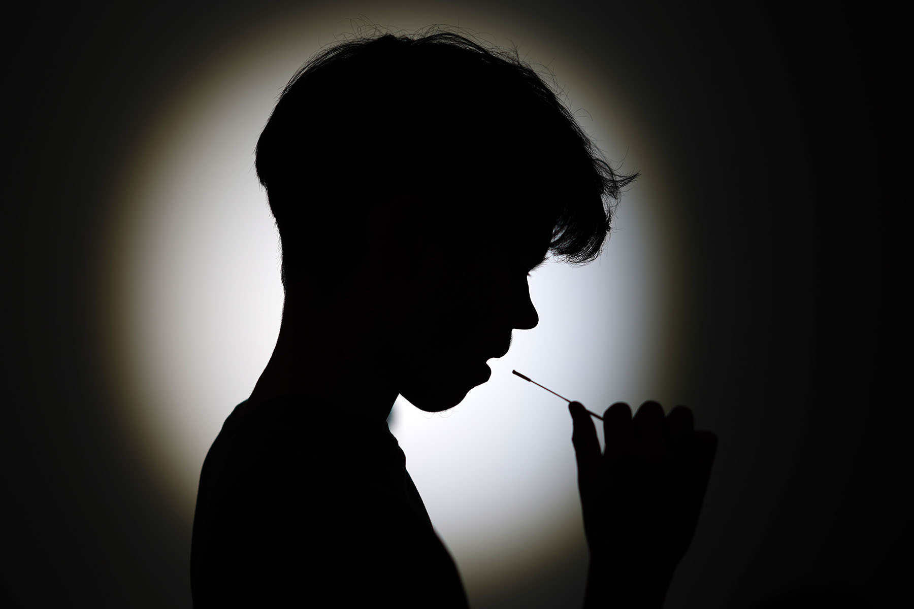 A silhouetted child holds a thin swab to their mouth, while taking an oral COVID-19 test against a softly lit background.