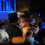 A toddler looks back down a street while held in the arms of an adult.