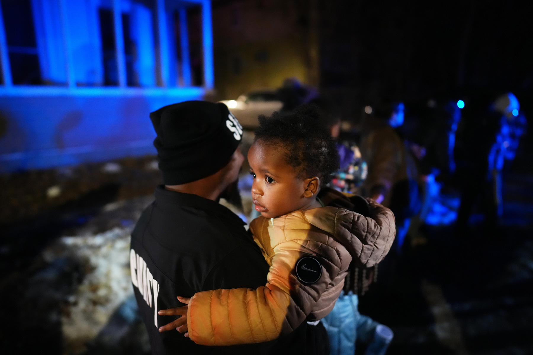 A toddler looks back down a street while held in the arms of an adult.