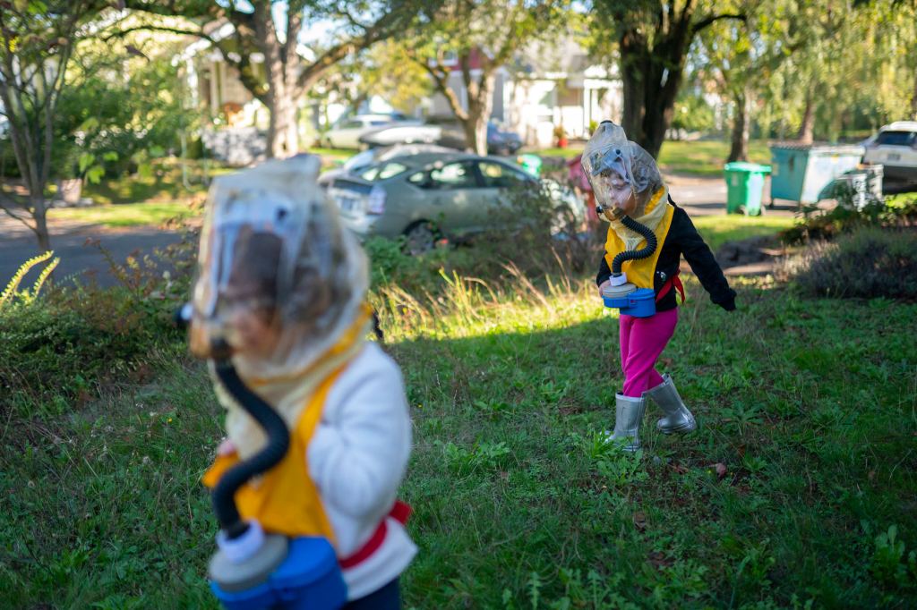 Two young children walk in a yard wearing gas masks.